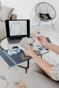 A woman reads tarot cards at a table with a laptop displaying an astrology chart in a modern living room setting.