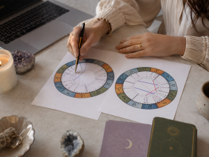 Hands reviewing two colorful natal charts with a pen beside a laptop, candle, crystals, and journals on a light stone desk.